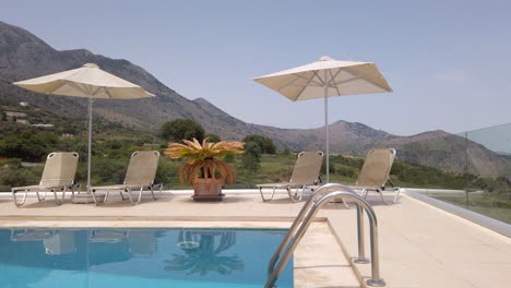 Gimbal-Aufnahme,-Die-Auf-Sonnenliegen-Auf-Der-Terrasse-Einer-Luxuriösen-Griechischen-Villa-Mit-Pool-Im-Vordergrund-Und-Bergen-Im-Hintergrund-Drückt