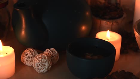 Relaxing-background-detail-shot-of-an-herbal-tea-on-a-green-cup,-with-steam-coming-out,-near-candles-with-flickering-flames,-a-tea-pot,-herbs-and-some-dust-flying-around