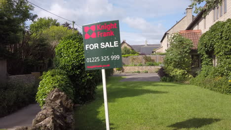 Left-to-Right-Truck-Shot-of-British-‘For-Sale’-Real-Estate-Sign-on-Sunny-Summer’s-Day-with-Rural-Village-Home-in-Background