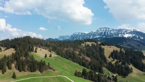 smooth-aerial-view-of-mountain-landscape-in-entlebuch,-switzerland,-spring-time-with-pine-trees-and-snowy-mountains-cloudy