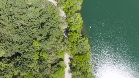 Birdseye-view-of-a-suv-hauling-two-jetskies-behind-driving-on-a-tight-forest-covered-road-next-to-a-beautiful-green-watered-lake-in-Wisconsin,-USA
