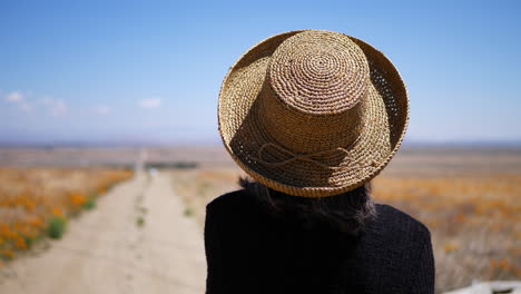 An-old-woman-in-summer-hat-looking-down-an-abandoned-dirt-road-in-a-field-of-orange-California-poppy-flowers-SLOW-MOTION