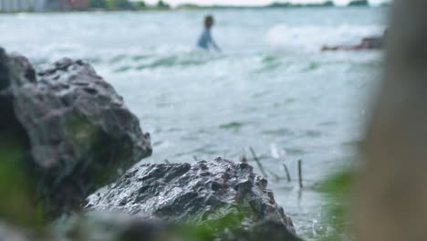 Wellen-Schlagen-Gegen-Felsen,-Während-Surfer-Im-Hintergrund-Zum-Wasser-Hinausgehen