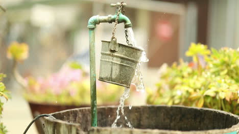 Water-from-a-garden-spout-pours-and-spills-over-a-hanging-bucket-into-a-larger-barrel