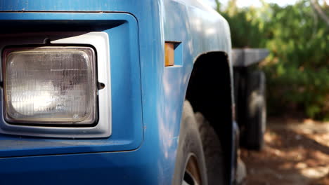 Slide-right-along-the-headlight-and-side-of-an-old-vintage-blue-cargo-truck-with-tires-parked-on-a-dirt-lot