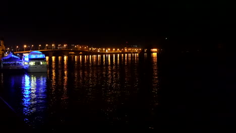 Beleuchtete-Brücke-über-Den-Fluss-Dnjepr-Bei-Nacht-Mit-Boot-Im-Vordergrund