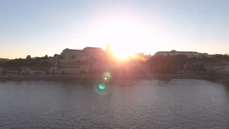 Static-shot-of-a-Drone-above-the-Danube-river,-sun-flairs-from-behind-the-castle-side-of-Buda