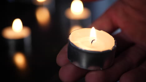 A-hand-holding-a-tealight-candle-on-fire-in-a-church-temple-with-other-candles-burning-and-heavy-background-blur-bokeh-behind