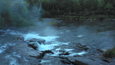 Aerial-drone-shot-over-a-dark-misty-stream-and-small-waterfalls-at-sunset-with-forest-trees-in-silhouette