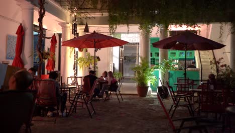 Gente-En-Mesas-Con-Sombrillas-Por-La-Noche-En-Un-Café-Restaurante-Que-Está-Situado-En-El-Patio-Central-De-Un-Gran-Edificio-Antiguo