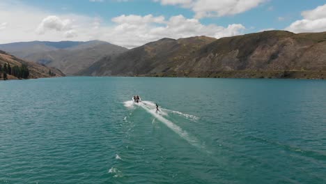 Cámara-Lenta:-Esquí-Acuático-Detrás-De-Un-Bote-En-El-Lago-Dunstan-Cerca-De-La-Represa-Clyde,-Otago-Central,-Nueva-Zelanda-Con-Montañas-Y-Nubes-En-El-Fondo