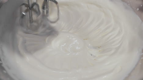 Close-up-macro-view-of-metal-mixer-stirring-and-blending-sweet-white-icing-in-circles-in-large-bowl-in-slow-motion