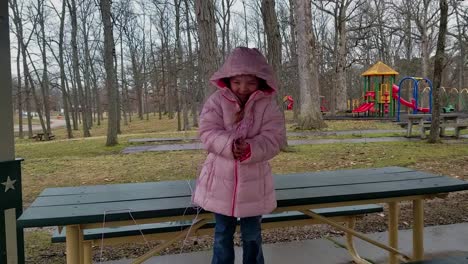A-young-girl-standing-on-a-picnic-table-in-a-pink-coat-at-the-playground-in-a-park
