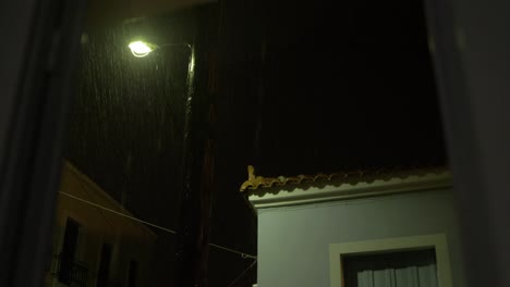 Lightning-lights-up-night-sky-during-storm-with-torrential-rainfall-around-streetlight-viewing-from-window-handheld