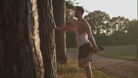 Man-In-Park-Stretching-Out-His-Legs-Before-He-Goes-For-a-Run-Whilst-Sun-Sets-Behind-Him-In-Slow-Motion