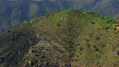 Un-Hermoso-Dron-Conduce-Sobre-La-Cima-De-La-Montaña-Verde-Cruzando-Las-Casas,-Otras-Hermosas-Montañas-En-El-Fondo