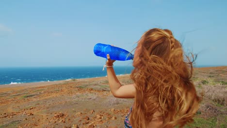 Beautiful-woman-drinking-from-blue-water-bottle-by-ocean-with-wind-blowing-hair