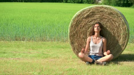 Sliding-right-across-an-attractive-brunette-woman-sitting-calmly-while-meditating-and-practicing-mindfulness-against-a-large-hay-bale-in-a-field-in-France