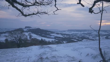 Timelapse-of-a-wintry-snowy-covered-English-farmland-scene-with-thicketed-branches-at-the-top-of-the-shot