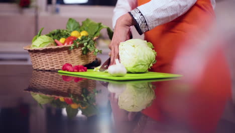 A-slow-mo-side-angle-close-up-view-of-a-lady-chef-cutting-cabbage-with-knife-in-the-kitchen,-A-basket-of-veggies-with-tomatoes-and-garlic-on-the-table,-The-chef-wore-and-orange-colored-apron