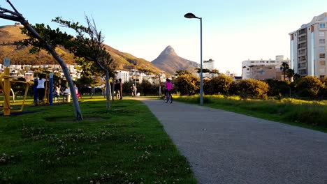 Green-Point-Park,-Recreation-area,-children-playing---girl-cycles-though-shot-with-views-out-to-South-Africa,-Cape-Town-Lions-Head-mountain-peak-along-the-skyline