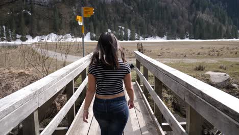 Junge-Asiatische-Frau-Mit-Langen-Haaren-Und-Gestreiftem-Hemd-Und-Jeans-Geht-In-Zeitlupe-über-Eine-Holzbrücke-In-Der-Schweiz
