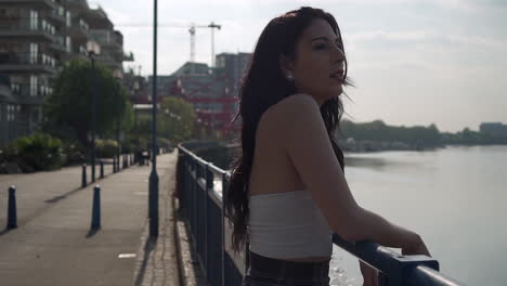 Beautiful-latina-woman-on-holiday-leaning-against-the-railing,-looking-at-the-river-Thames-in-London,-then-turning-at-the-camera,-smiling-and-wandering