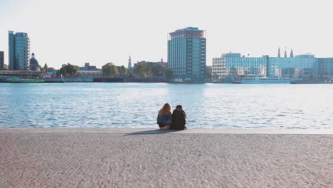 A-boy-and-a-girl-sitting-close-to-each-other-at-the-edge-of-a-dock-boulevard-with-the-river-and-cityscape-behind-them