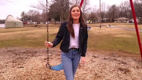 4K-footage-of-a-beautiful,-young,-brunette-college-teenager-standing-next-to-a-swing-set-in-a-city-park-talking-and-laughing