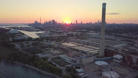 Aerial-Sunset-Wide-Shot-Flying-Over-Industrial-Factory-Past-Huge-Smokestack-Tower-With-Downtown-Skyline-In-Background-In-Toronto-Ontario-Canada