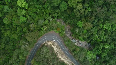 Aerial-view-of-a-car-driving-in-narrow-turning-road-in-the-middle-of-a-rainforest-with-nearby-waterfall-and-creek-flowing-with-water