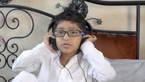 Cute-little-indian-asian-caucasian-boy-with-spectacles-listening-music-on-headphone-dancing-nodding-head-sitting-in-living-room-bedroom-looking-at-camera-front-view