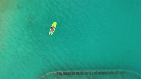 Birds-eye-view-of-woman-stand-up-paddle-over-rusty-shipwreck-in-clear-turquoise-water-by-tropical-island