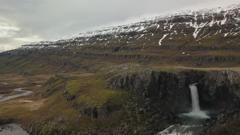 Cascada-Que-Fluye-Hacia-Un-Exuberante-Valle-Cubierto-De-Nieve-En-Islandia
