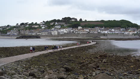 Tourists-walking-on-the-causeway-that-leads-to-Marazion-from-the-english-medioeval-castle-and-church-of-St-Michael's-Mount-in-Cornwall-on-a-cloudy-spring-day,-Zoom-out-4k-footage