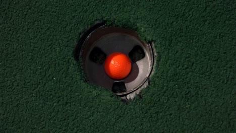 A-close-up-birds-eye-view-of-an-orange-mini-golf-ball-falls-into-the-golf-hole-and-bounces-in-the-pocket-on-a-course