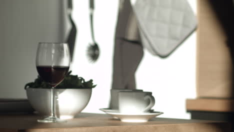 A-person-sets-out-two-coffee-cups-and-saucers-next-to-a-glass-of-wine-and-salad-bowl