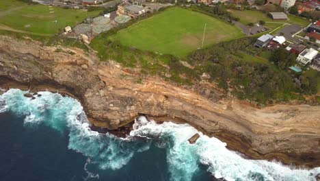 Epic-drone-shot-of-ocean-waves-smashing-against-cliff-and-rocks,-near-a-park,-Sydney,-Australia