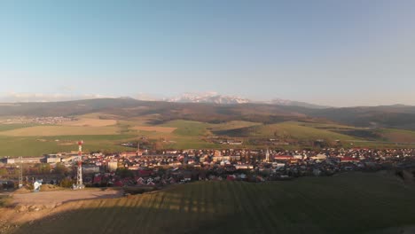 Forward-aerial-of-small-town-at-sunset-with-hills-and-snow-covered-mountains-in-background