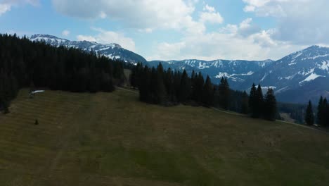 smooth-aerial-view-of-mountain-landscape-in-entlebuch,-switzerland,-spring-time-with-pine-trees-and-snowy-mountains-cloudy