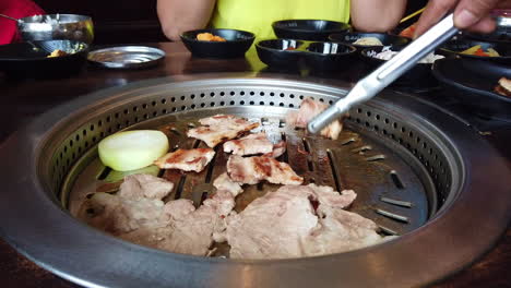 Eine-Nahaufnahme-Eines-Geschnittenen-Schweinefleischs-Auf-Einem-Koreanischen-Grill-Mit-Einer-Zwiebelscheibe-Und-Einer-Grillzange