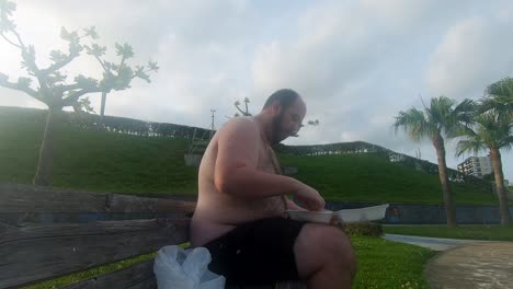 Obese-topless-man-exposes-his-fat,-beer-belly-while-eating-handfuls-of-chips-on-a-park-bench