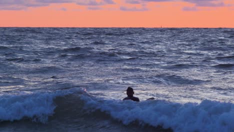 Surfer-on-surfboard-paddling-over-waves-near-the-Baltic-sea-Karosta-beach-at-Liepaja-during-a-beautiful-vibrant-sunset-at-golden-hour,-medium-shot-from-a-distance