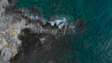 Aerial:-Drone-flying-down-to-fissured-cliffs-surrounded-by-waves-and-clear-water