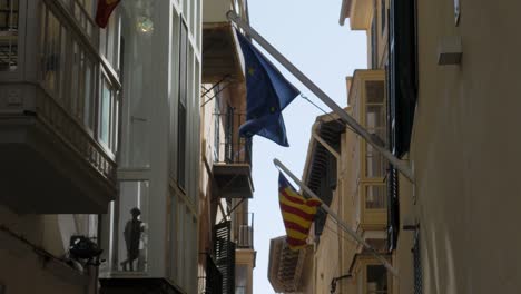 Europäische-Flagge-Und-Katalonien-Flagge-Winken-Auf-Einem-Fahnenmast,-Der-An-Einem-Gebäude-Unter-Einem-Fenster-In-Einer-Engen-Gasse-Befestigt-Ist