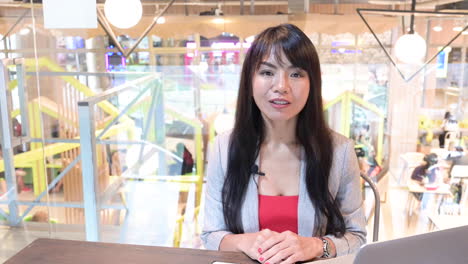 Schöne-Asiatische-YouTube-Frau,-Die-Vor-Der-Kamera-Mit-Einem-Laptop-Im-Arbeitszimmer-Spricht-Und-Im-Hintergrund-Arbeitende-Menschen