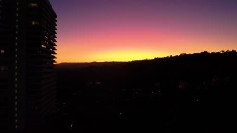 Climbing-higher-with-the-silhouette-of-a-high-rise-building-on-the-left,-as-the-sun-sets-behind-the-Hollywood-Hills