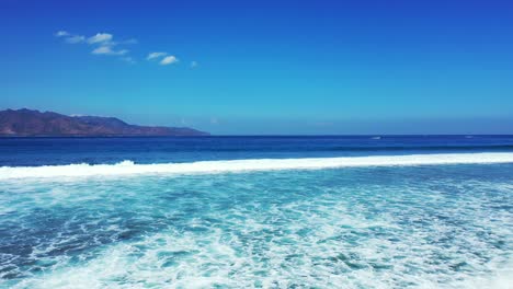 Die-Weißen-Wellen-Des-Tiefblauen-Ozeans-Plätschern-In-Richtung-Meeresküste-Im-Hintergrund-Mit-Hohen-Bergen-Und-Strahlend-Blauem-Himmel-–-Weitwinkelaufnahme
