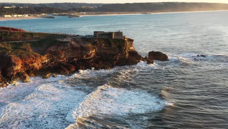 Der-Leuchtturm-Von-Nazare-Portugal-In-Praia-Do-Norte-Während-Des-Sonnenuntergangs-Zur-Goldenen-Stunde,-Luftaufnahme-Von-Der-Seite
