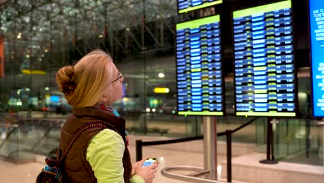 Side-shot-of-blond-woman-pointing-to-flight-information-display-while-holding-her-smartphone-in-an-airport